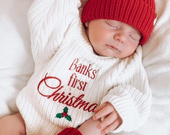Puede incluir: Un bebé dormido con un body de punto blanco con el texto bordado "Banks' first Christmas" en rojo. El bebé también lleva un gorro de punto rojo con un pompón blanco y botines de punto rojo. Una taza roja con crema batida está en el fondo.