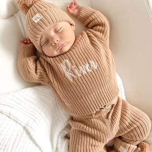 May include: A tan knitted baby outfit set, including a beanie with a pom-pom, a sweater with the name "River" embroidered, pants, and booties. The baby is lying on a white surface.