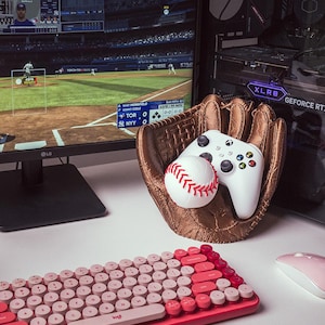Op de afbeelding: Een roze en rood retro-toetsenbord met ronde toetsen, een witte honkbal en een witte Xbox-controller in een bruine honkbalhandschoen. De handschoen ligt op een wit oppervlak. De tekst "log" is zichtbaar op het toetsenbord.