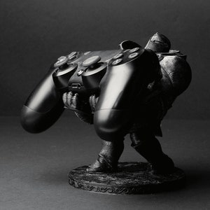 May include: A black and gray 3D printed figurine of a person holding a black PlayStation controller. The figurine is standing on a round base with a decorative design.