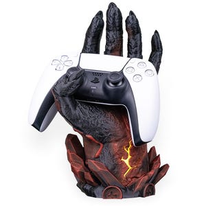 Può includere: Un controller PlayStation 5 bianco e nero tenuto da un supporto a forma di mano nero e strutturato. Il supporto ha una base rossa e nera con un design giallo brillante.