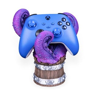 Op de afbeelding: Een blauwe Xbox-controller rust op een houten vat, met paarse tentakels die eruit komen en eromheen wikkelen. Het vat heeft zilveren metalen banden en studs. Een gaming accessoire.