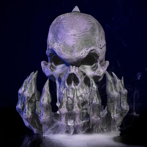 Puede incluir: Una detallada escultura de calavera gris con una expresión amenazante, rodeada de manos estilizadas. La escultura está parcialmente oscurecida por una nube de vapor blanco, sobre un fondo azul oscuro. La calavera tiene una superficie texturizada.