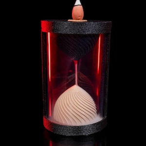 Puede incluir: Un porta incienso con forma de reloj de arena con base y parte superior negras. El soporte presenta una carcasa cilíndrica transparente con iluminación interna roja. En el interior, se ve una parte superior negra en forma de espiral y una parte inferior crema en forma de espiral. Encima hay un pequeño cono de incienso.