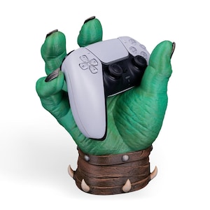 Könnte beinhalten: Eine grüne Monsterhand, die einen weiß-schwarzen Videospiel-Controller hält. Die Hand hat lange schwarze Fingernägel und ein braunes Lederarmband mit Metallnieten und Stoßzähnen. Der Controller hat ein modernes Design.