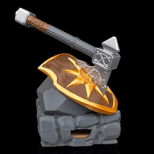 Könnte beinhalten: Ein Miniatur-Fantasy-Axt- und Schild-Set. Die Axt hat einen silbernen Kopf und einen braunen und grauen Griff. Das Schild ist braun mit einem goldenen Sonnenstrahl-Design. Das Set ruht auf einem grauen Steinsockel.