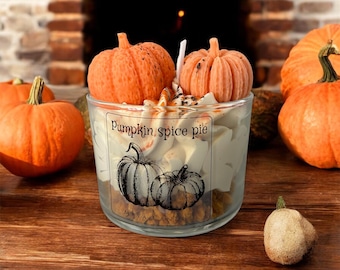 May include: A clear glass candle with a label that reads "Pumpkin Spice Pie" and features two pumpkin illustrations. The candle is topped with two small orange pumpkin-shaped decorations. Several pumpkins are arranged around the candle on a wooden surface.