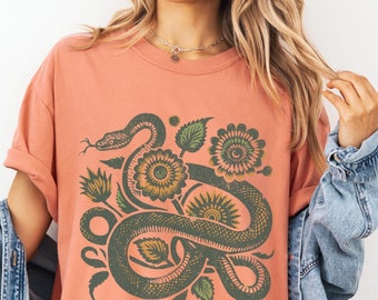 Könnte beinhalten: Ein pfirsichfarbenes T-Shirt mit einem grafischen Aufdruck einer grünen Schlange, die sich durch ein Blumenmuster mit Sonnenblumen windet.