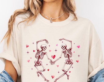 May include: A white t-shirt with a graphic of two skeletons dancing with pink hearts and stars.