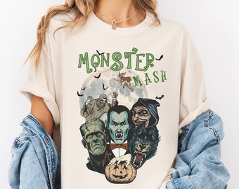May include: A cream-coloured t-shirt with a graphic of classic Halloween monsters, including Frankenstein's monster, Dracula, and a werewolf, with the text "Monster Mash" in green above them. The design is set against a full moon backdrop.