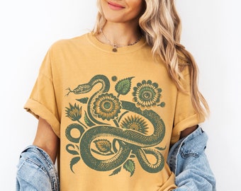 Könnte beinhalten: Ein senfgelbes T-Shirt mit einer grünen Schlange und einem floralen Design. Die Schlange ist um eine Sonnenblume und andere Blumen gewickelt.