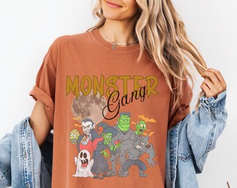 May include: A rust-coloured t-shirt featuring a graphic print with the text "MONSTER Gang" above a full moon and a group of cartoon monsters including a ghost, witch, vampire, werewolf, and Frankenstein's monster against a background of bats.