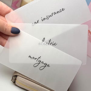 May include: A hand holds three translucent white cards with black script: "car insurance", "electric", and "mortgage". The cards are above a small, beige zippered wallet. The background is blurred.