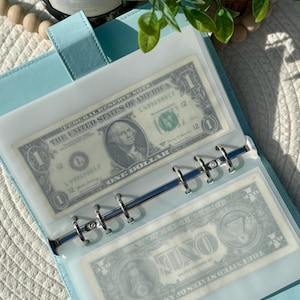 May include: A pale blue binder with silver rings, containing two one-dollar bills. The bills are visible through clear plastic sleeves. The binder is open, showing the bills and the interior design.