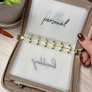 May include: A taupe-colored zippered wallet with a gold-tone ring binder. The wallet contains clear plastic inserts, one labeled "personal" in black script. A small zippered pocket is visible inside the wallet.