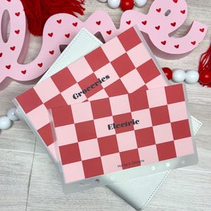 May include: Pink and red checkered budget envelopes labeled "Groceries" and "Electric" with a white wallet. The envelopes have a clear plastic border and the text "Honey b Designs". Decorative pink "Love" sign with red hearts.