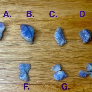 May include: Eight blue-violet rough gemstones are arranged on a wooden surface, labeled A through H. The stones vary in shape and size, with a mix of rounded and angular forms. The gemstones have a translucent quality, with some showing lighter, almost white, areas.