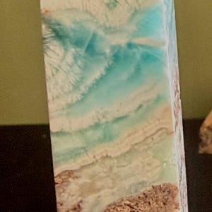 May include: A rectangular, polished stone obelisk with a flat top. The stone features layers of beige, brown, and turquoise hues, creating a natural, textured appearance. The obelisk is a decorative item.