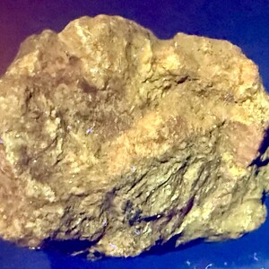 May include: A raw, irregularly shaped mineral specimen with a textured, earthy surface. The rock displays a muted golden-brown and olive-green coloration with visible crystalline facets and rough, jagged edges throughout its entire structure.