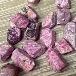 May include: A selection of rough ruby gemstones in shades of pink and red. The unpolished stones display natural textures and inclusions, with some showing dark spots. The gems are scattered on a light brown surface, creating a natural display.