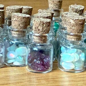 May include: A collection of small glass bottles filled with colorful gemstones and crystals. Each bottle is sealed with a cork stopper. The gemstones are various colors, including blue, purple, green, and clear.