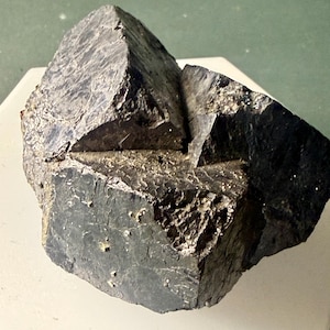 May include: A dark gray, metallic mineral specimen featuring sharp, angular, cubic crystal formations. The surface displays a rough, textured appearance with reflective facets and deep shadows, showcasing a complex, multi-faceted geometric structure.