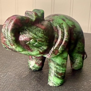 May include: A carved stone elephant figurine in shades of green and burgundy. The elephant has a trunk, four legs, and a small rectangular shape on its back. The stone has a polished finish.