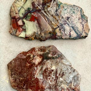 May include: Two irregularly shaped polished stones. The top stone features a mix of colors including red, green, white, and brown. The bottom stone is primarily red and brown with white and gray accents. Both stones have a natural, textured appearance.