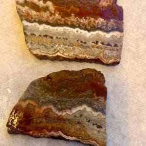 May include: Two polished, irregularly shaped stone slabs. The stones display layers of color, including shades of brown, red, white, and gray. The natural patterns and textures are visible, creating a unique and organic appearance. These stones could be used for decorative purposes.