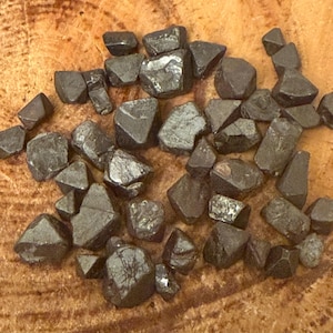 Pode incluir: Uma coleção de cristais de forma octaédrica, de cor cinza escuro, formados naturalmente. Os cristais estão dispostos sobre uma superfície de madeira.