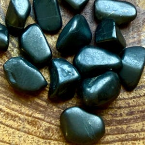 May include: A selection of smooth, dark green tumbled stones. The stones are polished and reflect light, with varying shapes and sizes. They are displayed on a textured, light brown wooden surface, giving a natural, earthy feel.