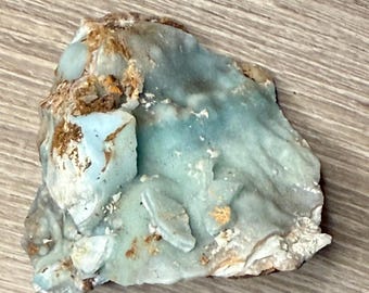 May include: A rough-cut, natural gemstone with a light blue hue, accented by areas of brown and white. The stone has a textured surface and irregular shape, showcasing its raw, unpolished state. This unique piece is ideal for collectors or jewelry makers.