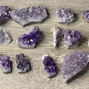 May include: A collection of amethyst crystal clusters in various shapes and sizes. The crystals display a range of purple hues, from light lavender to deep violet, with some having a crystalline structure. The pieces are arranged on a wooden surface.