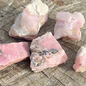May include: Five pink and white rough opal stones on a wooden surface. The stones are various sizes and shapes.