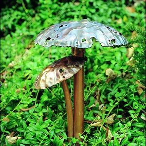 May include: Two copper mushroom sculptures with a textured surface and holes throughout. The sculptures are standing on a bed of green leaves.