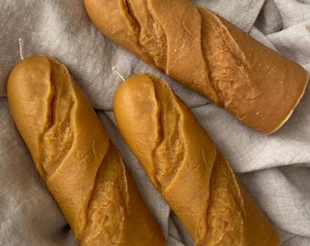 May include: Three long, brown, baguette-shaped candles with a textured surface. The candles are arranged on a light beige fabric.