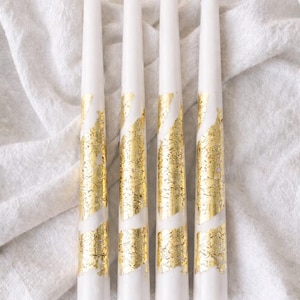 May include: Four white taper candles with gold leaf detailing. The gold leaf is arranged in a diagonal pattern around the candles. The candles are presented on a white, textured surface.
