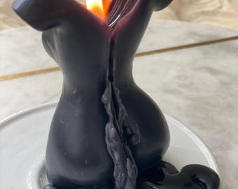 May include: A black candle shaped like a torso, with a lit flame at the top. The candle is on a white plate, with melted wax pooling around the base. The flame is bright orange and yellow.