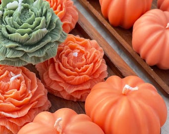 May include: A collection of decorative candles in various shapes and colors. The image features orange pumpkin-shaped candles, green and orange flower-shaped candles, and white pumpkin candles, all arranged on wooden trays. Each candle has a white wick.