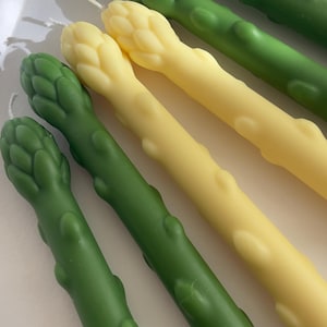 Puede incluir: Primer plano de velas con forma de espárragos en tonos verdes y amarillos. Las velas tienen una textura realista y una mecha en la parte superior. Las velas están dispuestas en diagonal, creando una exhibición visualmente atractiva.