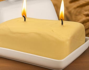May include: A yellow, rectangular candle with two wicks lit, placed on a white rectangular dish. A white lid is behind the candle. The background is blurred, with a wooden surface in the foreground.