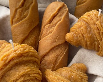 May include: A collection of six handmade candles shaped like bread and croissants. The candles are a light brown color and have a realistic texture.