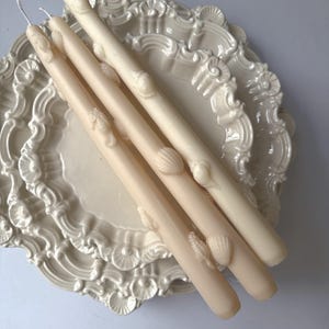 May include: Four ivory-colored taper candles with seashell and seahorse embellishments rest on a decorative, off-white plate. The candles vary slightly in shade, creating a subtle contrast. The plate has an ornate, scalloped edge.