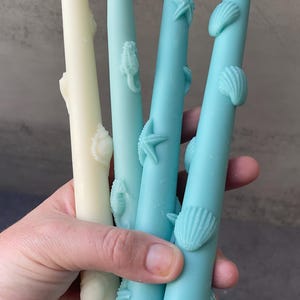 May include: Four decorative taper candles in shades of cream and blue, adorned with seashell and seahorse embellishments. The candles are held in a hand, showcasing their detailed designs. These candles are approximately 8 inches tall.
