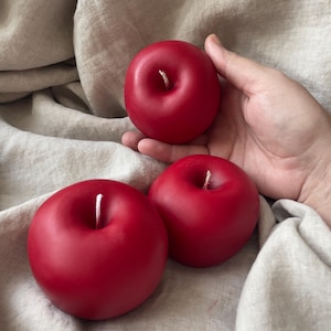 May include: Three red apple-shaped candles with white wicks. The candles are on a light brown fabric surface.