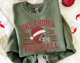 May include: An olive green sweatshirt with the words "OKLAHOMA FOOTBALL" in red lettering. A red football helmet with a Santa hat is in the center. The numbers "18" and "31" are on either side of the helmet. Two red Christmas ornaments are in the background.