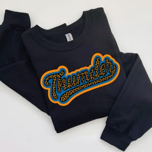 Op de afbeelding: Zwart sweatshirt met het woord "Thunder" in een retro-ontwerp. Het woord heeft een luipaardprintpatroon met een blauwe omtrek en een oranje rand. Het sweatshirt is op een witte ondergrond gevouwen.