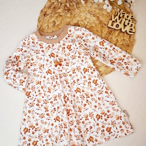 Könnte beinhalten: Weißes Langarmkleid mit floralem Muster in Braun- und Beigetönen. Das Kleid hat einen braunen Ausschnitt und eine kleine Schleife. Die Worte "Made with Love" sind auf einem Holzschild zu sehen.