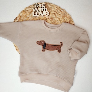 Könnte beinhalten: Beigefarbenes Waffelstrick-Sweatshirt mit einem braunen Dackel-Applikation. Das Sweatshirt hat einen Rundhalsausschnitt und lange Ärmel mit gerippten Bündchen. Die Worte "Made with Love" sind im Hintergrund zu sehen.