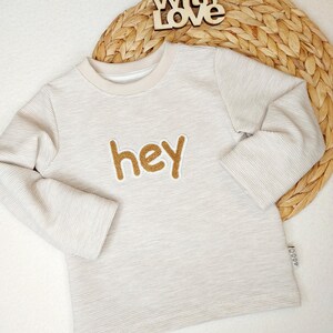 Könnte beinhalten: Ein hellbeige Langarmshirt mit dem Wort "hey" in goldglitzernden Buchstaben. Das Shirt hat eine gerippte Textur und einen beigen Rundhalsausschnitt. Ein Holzschild mit der Aufschrift "Made With Love" befindet sich im Hintergrund.